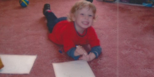 A photograph of toddler-aged Thomas, with blond hair, lying on a carpeted floor, clutching a pen and smiling gleefully.